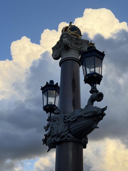 This photo captures an ornate street lamp, adorned with classical figures and a crown, standing tall against a dramatic sky filled with fluffy white and dark grey clouds